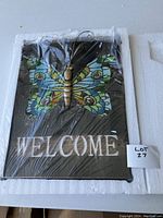 Front view of the solar butterfly welcome sign showing colorful butterfly and 'WELCOME' text on black plaque packaged in plastic.