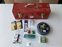 Red metal toolbox with assorted hand tools and hardware laid out in front on a white table.