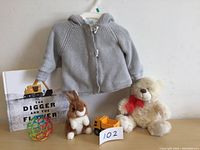 Front view of the lot showing the grey fleece lined hooded baby sweater, children's book, a colorful open ball toy, small plush bunny, toy construction vehicle, and cream teddy bear with red bow