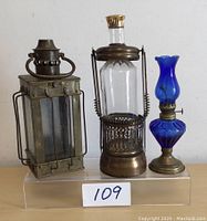 Front view of three items: rectangular brass lantern, brass and glass decanter with stopper, blue glass chimney on brass base