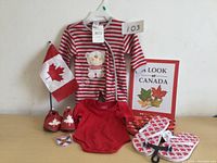 Front views of two baby sleepers (striped and solid red), two pairs of flip flops, baby slippers, Canadian flag, and book
