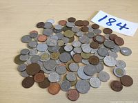 Overview of approximately 80 assorted foreign coins spread on a wooden surface showing a mix of copper, silver, and brass tones.