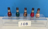 Five new bottles of Kylie nail polish in assorted shades displayed on a clear stand against a blue background with lot number 108 visible.