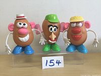 Three Mr Potato People toy figures with different hats and facial features arranged in a row on a clear platform providing front view of each figure.