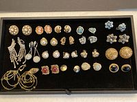 Full view of all 20 pairs of clip-on earrings arranged in a black lined tray, showing variety of materials and styles including pearls, blue stones, gold-tone filigree, and cameo designs.