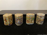 Four small cloisonné containers displayed in a row showing front designs