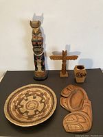 Five First Nations items displayed on a black surface: tall painted totem figure, smaller totem pole, carved bowl, woven basket, small painted wooden holder.