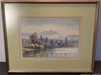 Framed watercolour painting showing a landscape of a river with trees and mountains under a cloudy sky.
