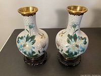 Front view of the pair of cloisonné vases showing detailed floral and bird enamel work and gold rims, each on a dark wood plinth.