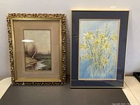 Photo showing both watercolour paintings side by side, one landscape with a gilt ornate frame and one floral still life with a dark frame and matting