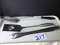 Three Napoleon BBQ utensils with long handles: grill brush, spatula, tongs with logo cutout and serrated edges.