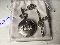 Front of Daniel David pocket watch showing raised train relief on cover along with attached chain on a white cloth pouch.