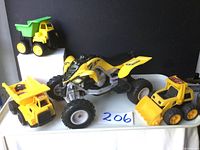 Four toy vehicles on a white surface against black background: yellow dump truck with green bed, yellow dump truck, yellow front end loader, yellow and black Yamaha quad bike