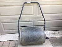 Side view of heavy plastic lawn roller against garage door, showing black metal handle and cylindrical roller surface.