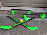Two street hockey sticks crossed with a yellow ball in front of two plastic hockey goals on pavement