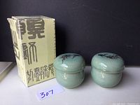 Two celadon glazed ceramic tea strainers displayed next to their original box with Korean text. Each strainer has a matching lid with floral design.