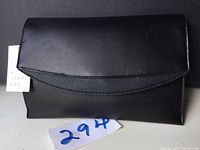 Front view of black handbag clutch with curved flap and pebbled accent strip, tag visible.