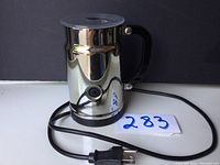 Front and side view of stainless steel electric milk frother with black handle and cord with plug visible.
