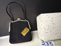 Black evening bag showing front and side profile with textured fabric and metal frame, accompanied by original box.