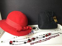 Red ladies hat, black handbag, three bead necklaces, and silver-tone bracelet on white surface against black background.