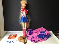 Full view of Sailor Moon doll standing upright with long blonde hair, wearing sailor outfit, next to folded pink umbrella with blue cat designs, brown stand, and three small sticker sheets.