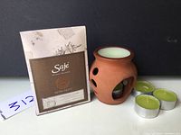 Photo showing the SAJE Soothing Bath Salt Soak box, clay ceramic candle warmer with white candle, and three green tea light candles.