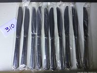 Ten stainless steel knives with black handles, arranged side by side, each individually wrapped in plastic.