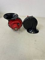 Two air horns on a white surface, showing one red and black horn and one black horn.
