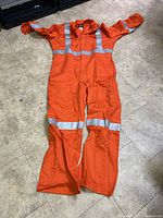 Front view of one men's orange coverall laid flat on tiled floor, showing reflective bands on chest, arms, and legs.