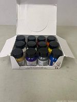 Top down view inside the box showing the arrangement of 12 bottles with black lids and labels featuring the Angelus brand.