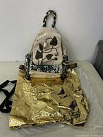 Full view of handwoven backpack with beige body, floral appliqué on front flap, embroidered pockets, and braided handles, displayed on a gold foil surface.