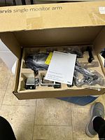 Top-down view inside the Jarvis single monitor arm box, showing black metal components, clamps, and an instruction manual labeled Fully.