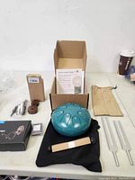 Photo of the teal handpan drum, black carrying pouch, wooden tuning tool, instructional material, headphones box, tuning fork striker, and small parts box opened with accessories.