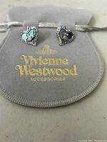 Pair of Vivienne Westwood earrings displayed on a grey dust bag with gold logo lettering. Earrings are heart-shaped with iridescent blue inlay and crystal border.