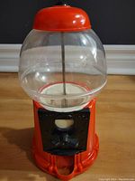 Front view of vintage red gumball machine on a wooden floor showing the clear globe, red base and black coin slot.