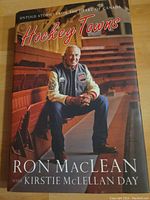 Front cover of 'Hockey Towns' book showing Ron MacLean seated in hockey arena bleachers.