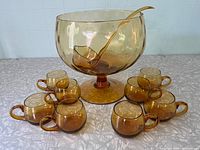 Full view of amber glass punch bowl with ladle inside surrounded by eight matching cups, displayed on white cloth.