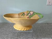 Side angle view of the oval bowl showing basket weave texture, floral handle with pink and purple flowers, and yellow rope-like pedestal base.