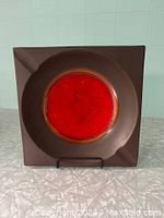 Front view showing square shape, red and orange circular design on brown ceramic ashtray.
