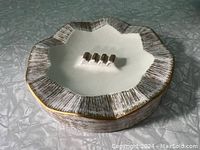 Top angled view of ceramic ashtray showing scalloped edges with textured white and gold pattern and three cigarette rests with gold accents, on a grey surface.