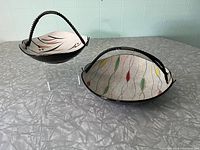 Two ceramic baskets with wire handles, showing colorful hand-painted wavy leaf designs on cream speckled background and black exteriors.