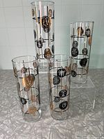 Four tall cylindrical highball glasses decorated with gold and black Roman medallion motifs on a light surface, showing all four glasses standing together