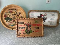Three kitschy bar trays displayed together: round Florida map tray, rectangular camel quote tray, and rectangular rooster recipe tray.