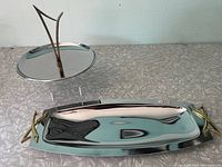 Photo showing both the round two-tier serving dish and rectangular serving tray on a textured silver surface against a tile wall.