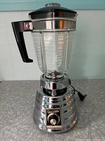 Full view of vintage Osterizer blender showing glass pitcher and chrome body with black handle and lid.