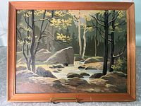 Paint by numbers landscape painting in wooden frame showing a forest scene with a river, rocks, and trees.