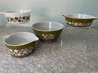 Four Pyrex Spring Blossom Green mixing bowls displayed on grey surface, two raised on clear stands. Olive green exterior with white floral pattern on three bowls, one bowl has white background with green flowers.