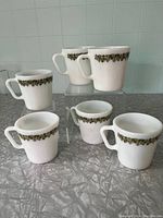 Six white milk glass coffee cups with green floral band pattern, arranged on a counter, showing overall set.