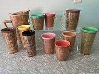 Full view of the eleven-piece Raffiaware collection including beige pitcher and various glasses and mugs with colored rims, all featuring woven raffia pattern exterior.