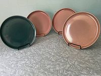 Four Russel Wright American Modern plates, three coral color and one seafoam green, displayed on stands on a tabletop.